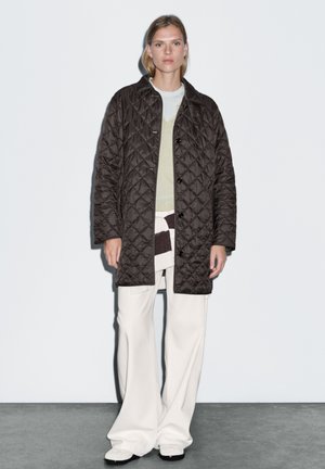 Quilted dark brown coat with button closure, worn over a light green shirt and cream wide-leg pants, featuring a textured surface.