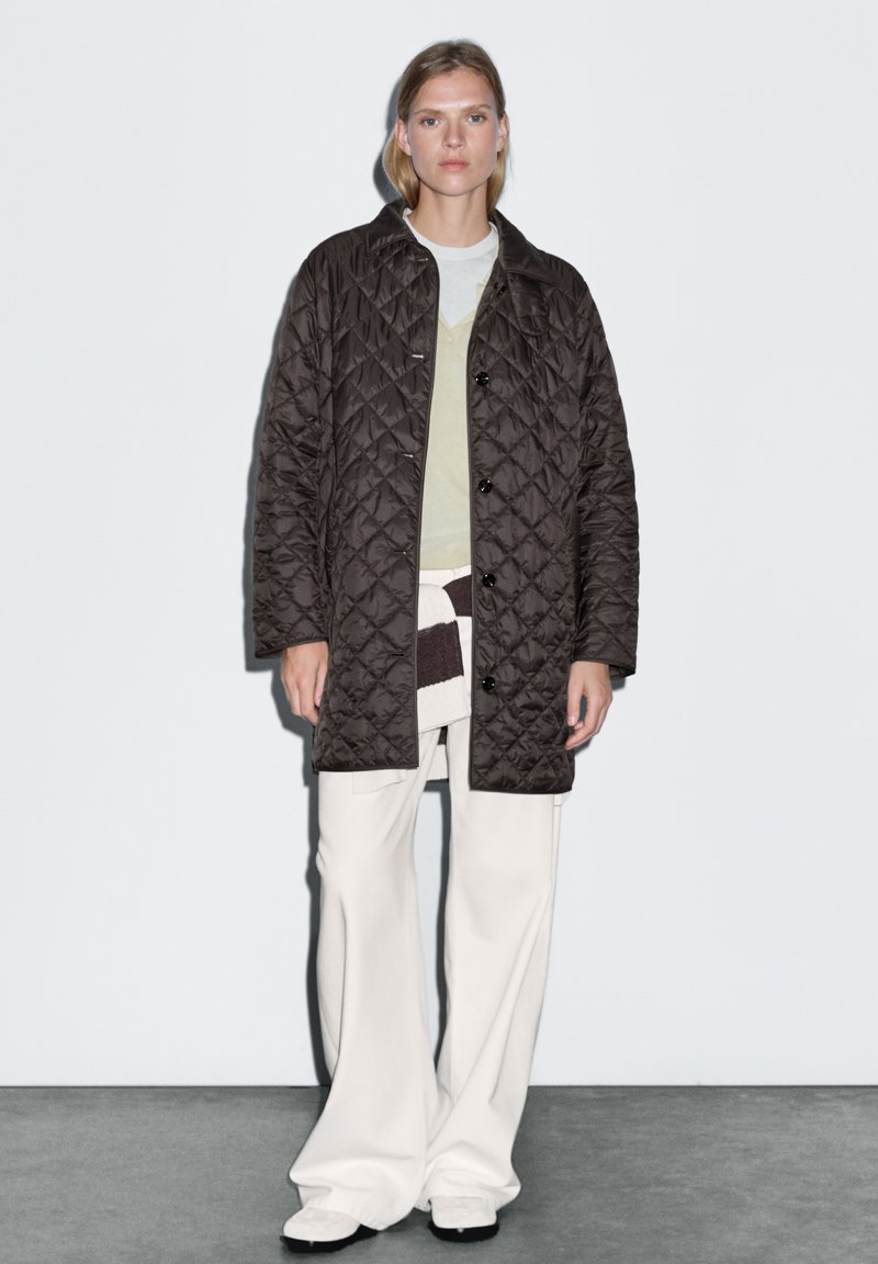 Prošívaná hnědá bunda s knoflíkovým zapínáním, nošená přes světle zelený top a krémové kalhoty s širokými nohavicemi, s různými texturami.