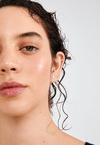 Artimiausiu planu pateiktas veidas su šlapiomis, garbanojomis šukuosenomis. Išryškintos mažos auksinės auskarai skirtingų apvalių dizainų ir lygi, šiek tiek blizgi odos tekstūra.