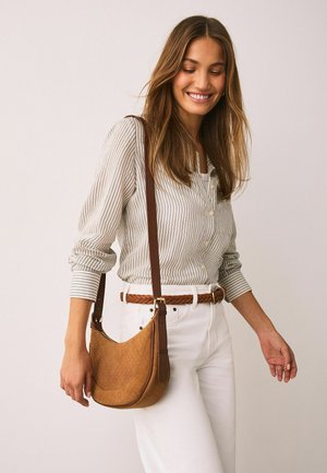 Brūna ādas plecu soma ar teksturētu, austu rakstu, ar garu siksnu un zelta toņa metāla detaļām, kombinēta ar svītrainu kreklu un baltām biksēm.