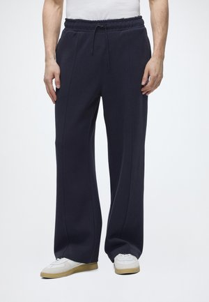 Unterkörper einer Person, die marineblaue, weit geschnittene Hose mit elastischem Bund und weiße Sneakers mit Gummisohlen trägt.
