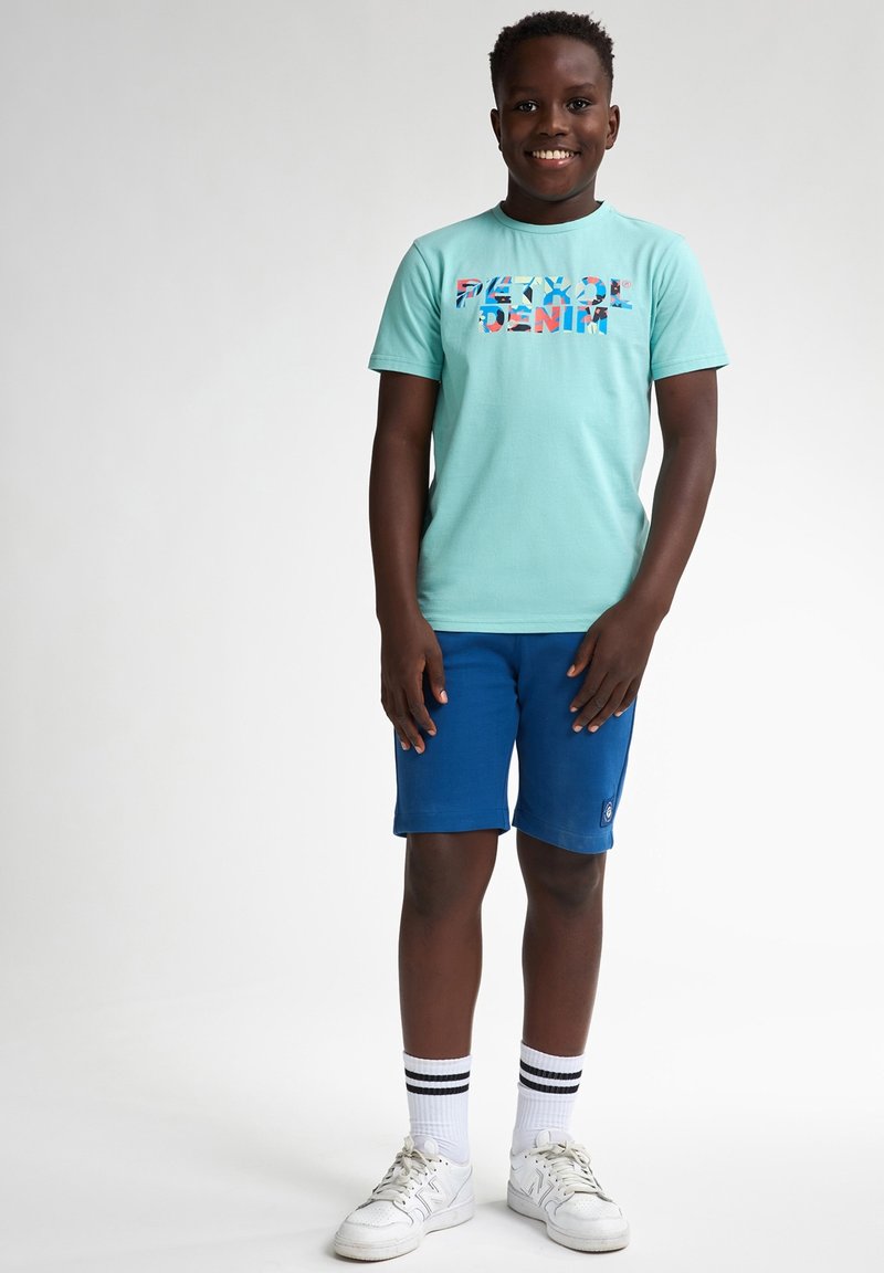 Lichtblauwe T-shirt met een kleurrijk grafisch tekstontwerp, gecombineerd met blauwe shorts. Het model draagt witte sneakers en gestreepte sokken.