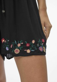 Vêtement en tissu noir avec une broderie florale colorée à l'ourlet, incorporant des teintes de rouge, rose, jaune et bleu, doté d'une surface texturée.