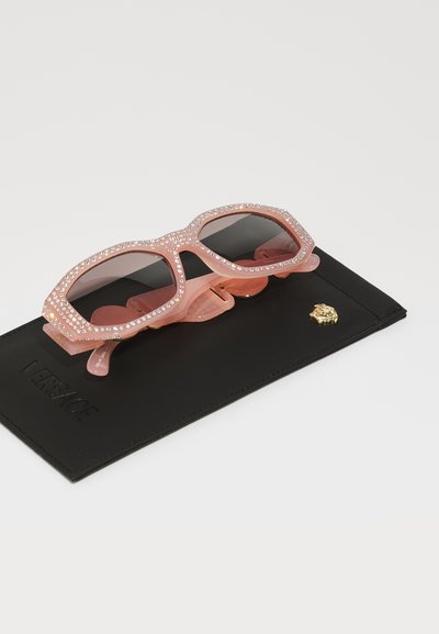 Óculos de sol oversized cor-de-rosa com lentes pretas, adornados com strass ao longo da armação, repousando sobre uma caixa de couro preto com logotipo embossado.