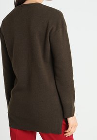 Donna che indossa un maglione a maglia a maniche lunghe di colore marrone scuro e pantaloni rossi, vista di spalle contro uno sfondo chiaro e semplice.
