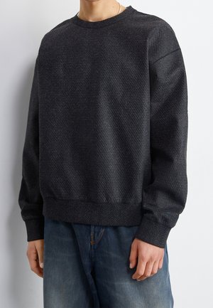 Person trägt einen schwarzen, strukturierten Langarmpullover und blaue Jeans und steht vor einem einfachen weißen Hintergrund.