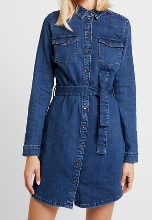 Woman wearing a dark blue denim shirt dress with long sleeves, button-front closure, chest pockets, and a waist tie belt.