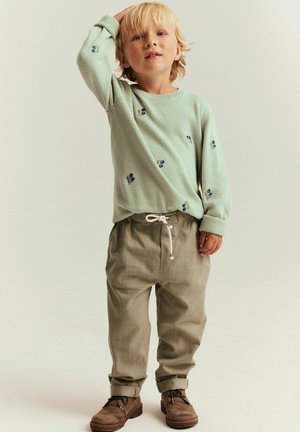 Enfant blond se tenant la tête d'une main, portant un pull vert clair avec de petites fleurs bleues imprimées, un pantalon beige et des chaussures marron.