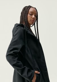 Črna kapucinska jakna z gladko teksturo, sprednjimi gumbi in stranskimi žepi. Oblikovanje vsebuje sproščen kroj in rahlo sijaj.