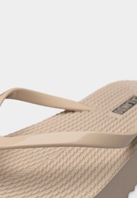 Gros plan sur une tong beige avec une semelle texturée et de simples lanières en caoutchouc, montrant une partie du dessus du pied et le profil latéral.