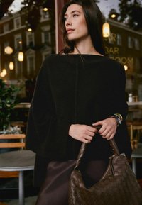 Woman with long dark hair wears a black cape, holds a large woven brown handbag, standing near a café window at dusk with reflections and lights.