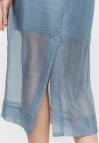 Blå genomskinlig klänning med ett lager på lager-design, som har en delad nederkant och ett lätt, texturerat tyg. Inkluderar en subtil transparent sektion.