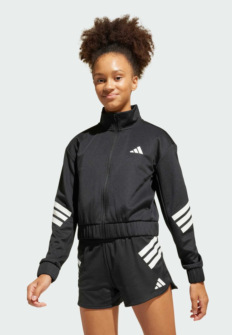 Giovane donna che indossa una giacca da allenamento nera Adidas e pantaloni corti coordinati con strisce bianche, in piedi contro uno sfondo chiaro e semplice.