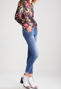Blouse à manches longues florale noire avec des fleurs roses et jaunes, associée à un jean skinny bleu clair et des bottines blanches à talons ouvertes aux orteils.