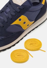 Navy kék sneaker sárga kiegészítőkkel, textúrált velúr és nylon anyagból, lekerekített orral, lapos cipőfűzőkkel és két sárga pótfűzővel.