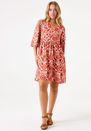 Robe à motifs rouge et blanc avec un décolleté en V, des manches courtes et une taille cintrée, ornée de motifs floraux et géométriques. Sandales à talons marron.