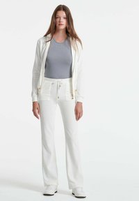 Bijela zip-up dukserica preko sive rebraste majice bez rukava, u kombinaciji s bijelim hlačama s vezicom koje imaju prednje džepove i zlatne detalje.