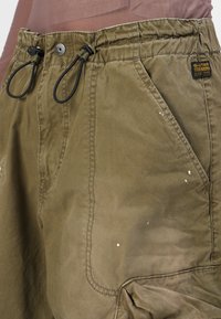Olivenfargede cargo-shorts med snøring i midjen, med frontlommer, stikninger og små hvite malingssprut.