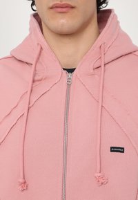 Hanorac roz cu fermoar, având o textură inegală, cusături laterale, șnururi cu accente din pompoane și un mic eticheta cu logo-ul "BLUEMARBLE".