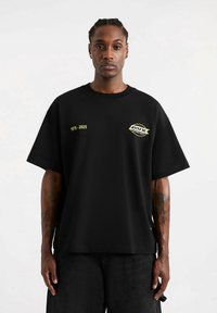 Czarna bawełniana koszulka z luźnym krojem. Posiada żółty napis "1975-2025" oraz logo na lewej piersi. Krótkie rękawy i okrągły dekolt.