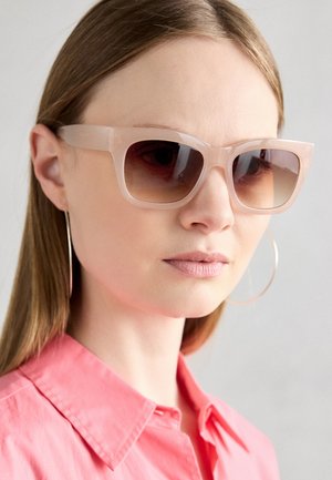 Translucente rosa solbriller med brede, vinklede rammer og gradient brune linser. Modellen har på seg hoop øredobber og en rosa skjorte med krage.