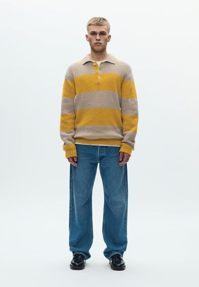 Πλεκτό πουλόβερ πόλο σε μπεζ με κίτρινες και γκρι ρίγες, με γιακά και τρία κουμπιά. Συνδυάζεται με άνετο μπλε τζιν.