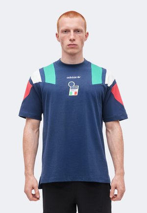Mladý muž s krátkymi červenými vlasmi, ktorý má na sebe tmavomodré tričko Adidas so zelenými, bielymi a červenými akcentmi a emblémom talianskej vlajky.