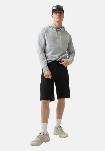 Grauer Hoodie mit neongrünem Text, schwarze Shorts und cremefarbene Sneakers. Das Outfit hat einen lässigen Schnitt und ein sportliches Design mit sichtbarem Branding.