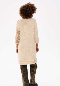 Gebreide jurk in lichtbeige met een textuur gebreid lijf en mouwen met luipaardpatroon, gecombineerd met luipaardafdruk broeken en zwarte schoenen.