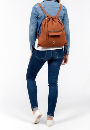 Femme portant une veste en jean bleue, un jean slim, des chaussures blanches, et un sac à dos marron à cordon avec une poche zippée à l'avant.