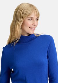 Trikós kék garbó pulóver bordás nyakkal és hosszú ujjal. Az anyag simának és testhezállónak tűnik, minimális varrásokkal és díszítéssel.