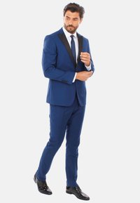 Completo blu con rever nero, design slim fit. Camicia bianca e cravatta, scarpe nere lucide. Modello che sistema il polsino, sfondo neutro.