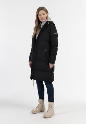 Vrouw draagt een zwarte doorgestikte lange jas met capuchon, blauwe spijkerbroek, beige veterschoenen en een lichtgrijze trui eronder.