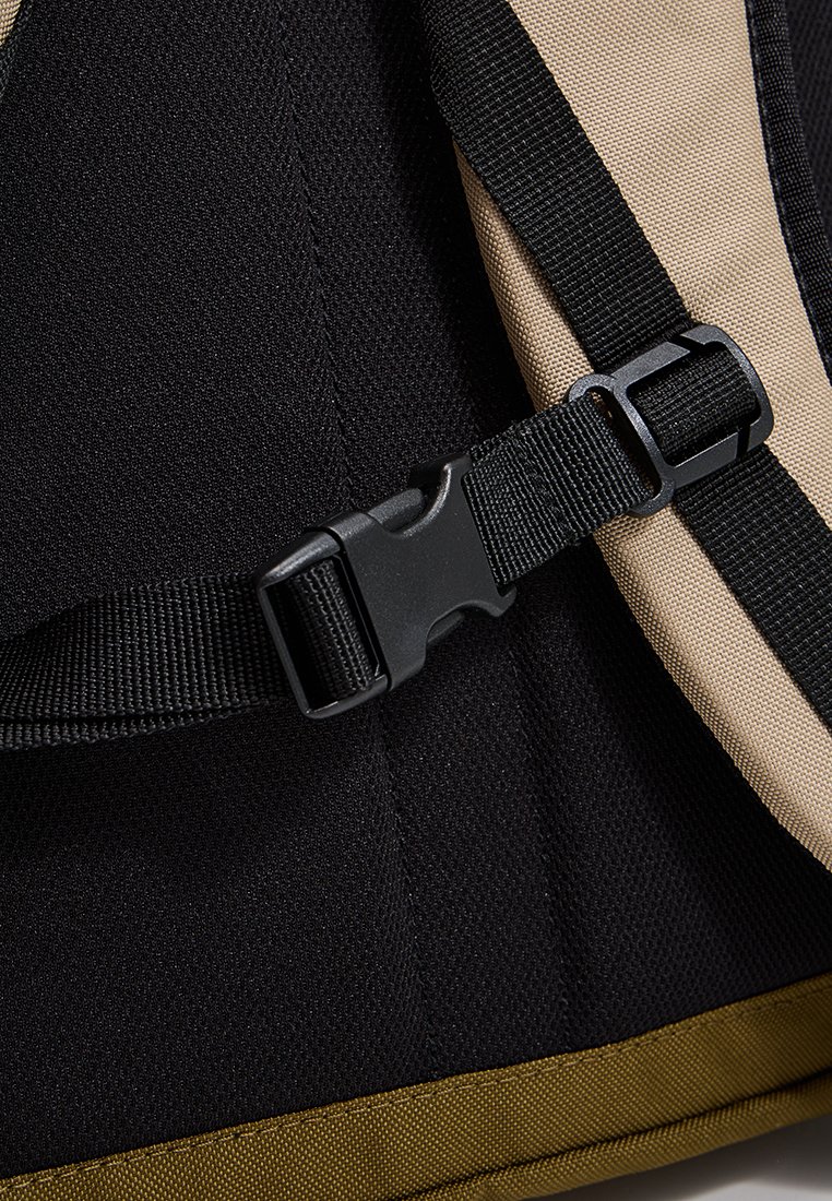 Eckige schwarze und beige Stoff-Rucksack mit einem stabilen Plastikverschluss und gewebten Trägern, der strukturierte Oberflächen und kontrastierende Farben aufweist.