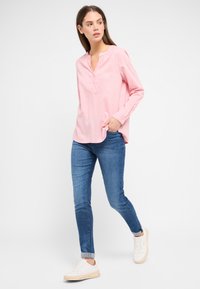Hellrosa Langarmshirt mit Stehkragen, ausgestattet mit einer Knopfleiste und kleinen weißen Punkten, kombiniert mit blauen Skinny Jeans und weißen Sneakers.