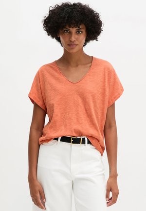 Kobieta z kręconymi włosami, w pomarańczowej koszulce z krótkim rękawem i dekoltem w serek, wsadzonej w białe spodnie z czarnym paskiem, stojąca na jednolitym tle.