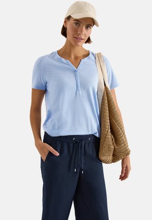 Kobieta w jasnoniebieskim topie z krótkim rękawem, granatowych spodniach ze sznurkiem, beżowej czapce, niosąca brązową plecioną torbę na ramię.