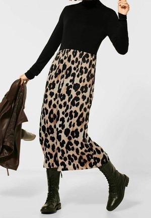 Osoba oblečená v černém tričku s dlouhým rukávem, leopardí sukni, černých šněrovacích botách, držící v jedné ruce hnědou koženou bundu.