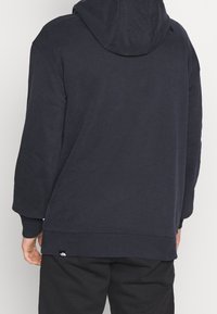 Námořnicky modrá mikina s kapucí a volnějším střihem, s žebrovanými lemy rukávů a spodního okraje. Na spodním lemu malá logo cedulka. Měkká textura látky.