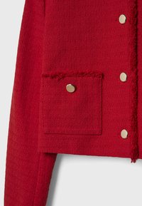 Manche et devant de veste en tissu rouge texturé avec une poche carrée dotée d'un seul bouton métallique et des boutons-pression métalliques le long du bord.