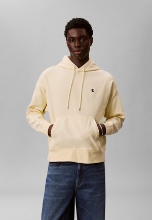 Young man wearing a light beige Calvin Klein hoodie with hands in the front pocket and blue jeans, standing against a gray background.