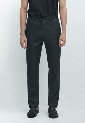 Uomo che indossa pantaloni neri aderenti, cintura nera e mocassini in pelle neri, in piedi davanti a uno sfondo grigio chiaro uniforme.