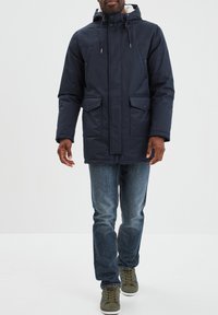 Parka con cappuccio di colore blu navy realizzata in tessuto con texture liscia, dotata di tasche frontali e chiusura con zip. Abbinata a jeans blu e scarpe da ginnastica verdi.