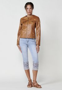 Giubbotto di pelle marrone con chiusura lampo e due tasche sul petto, abbinato a capri in denim azzurro chiaro con intricate ricami viola ai polsini.