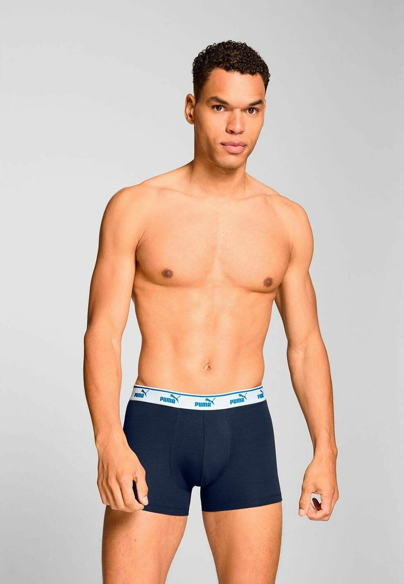 Man with short curly hair wearing navy blue Puma boxer briefs with white waistband, standing against a plain light gray background.