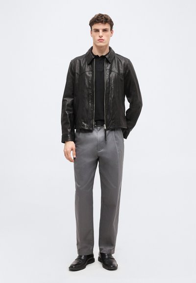 Homme debout portant une veste en cuir noire, une chemise noire, un pantalon gris et des chaussures noires sur un fond gris clair uni.