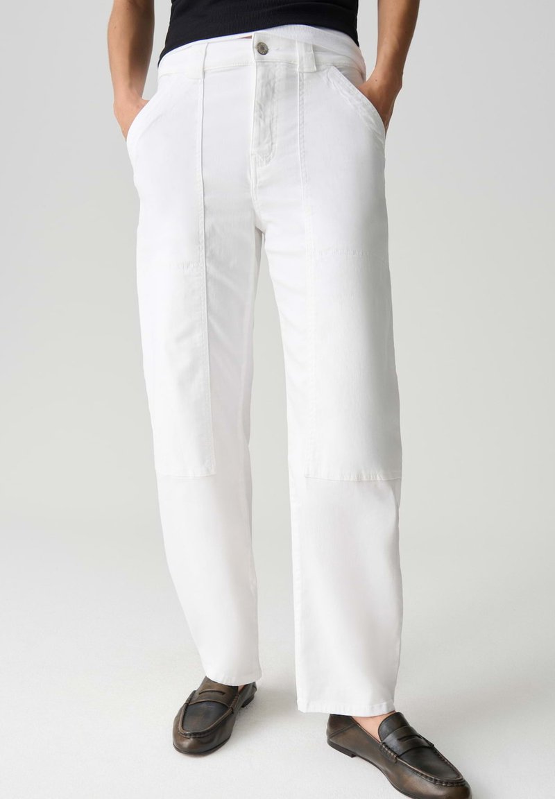 Persoon die witte, rechte broek met frontpatchdetails en donkerbruine loafers draagt, staand met handen in de zakken.