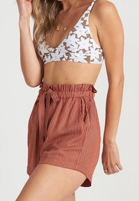 Bralette marrone con stampa floreale di fiori bianchi, abbinato a pantaloncini a righe ruggine a vita alta con cintura annodata e vestibilità morbida.