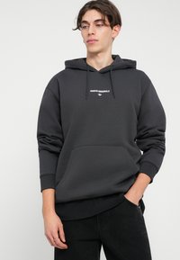 Tumši pelēks kapuci ar priekšējo kabatu, aukliņu kapuci un baltu "ADIDAS ORIGINALS" logo. Audums izskatās mīksts, ar brīvu piegriezumu.