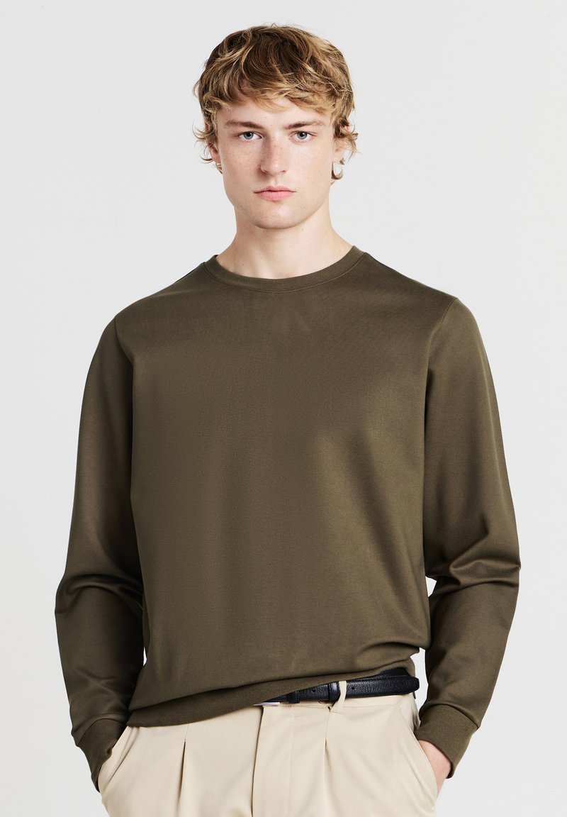 Camicia a maniche lunghe di colore verde oliva scuro, realizzata in tessuto liscio con collo a giro. Abbinata a pantaloni di colore chiaro e una cintura nera.